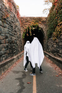 Two people dressed as ghosts walk hand in hand through a dim tunnel lit by overhead lights and covered in ivy.