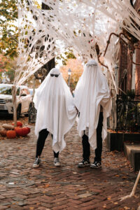 A couple dressed as ghosts walks toward Halloween decorations with arms raised playfully
