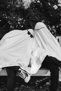 A ghost-costumed couple sits a bench, draped in white sheets and sunglasses with their heads leaning together.
