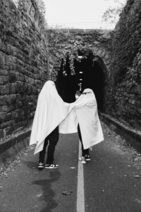 A couple in ghost sheets dance through a narrow tunnel surrounded by tall stone walls.